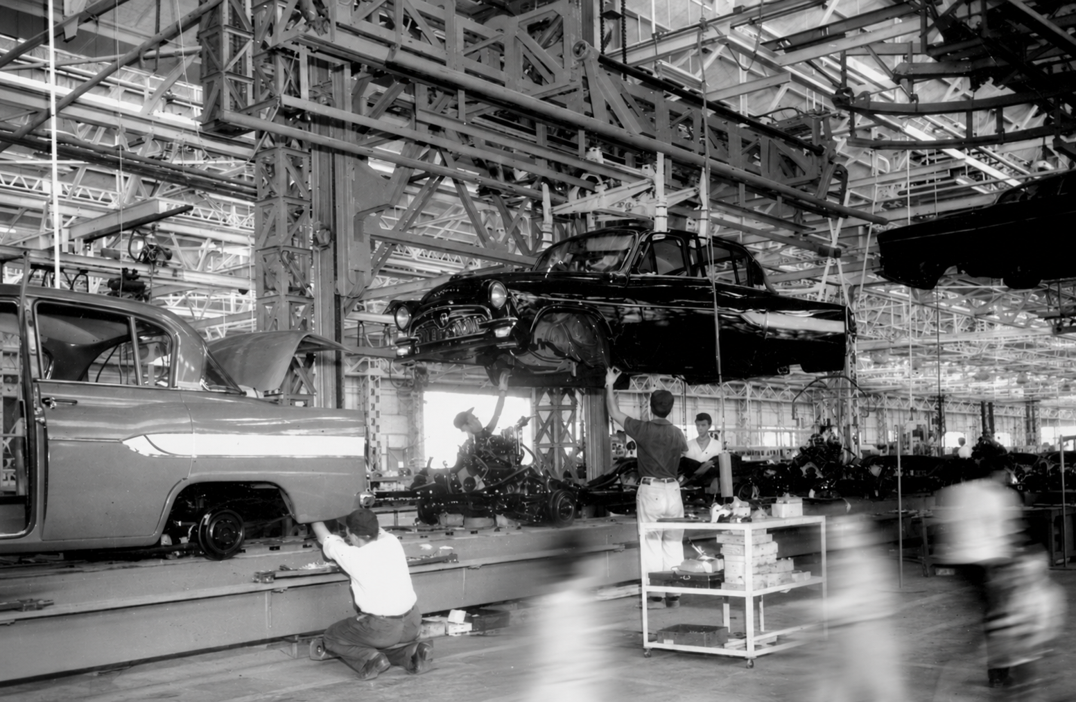Workers on a vintage Toyota assembly line, hand-fitting body panels to a sedan.